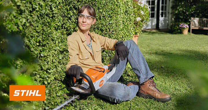 Frau lehnt an Hecke und hält STIHL Kettensäge, STIHL Logo sichtbar.