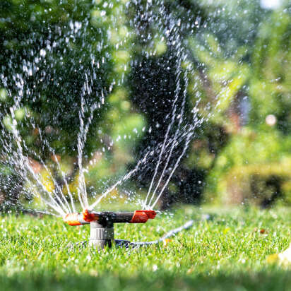 Sprinkler sprüht Wasser auf grünes Gras im Sonnenschein, umgeben von verschwommenen Blättern.